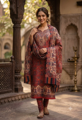 A woman models a traditional Indian salwar kameez suit set with an intricate red, gold, and blue floral pattern. The set includes a long kurta, matching trousers, and a draped dupatta, set against an outdoor architectural background with stone pillars and arches.