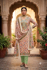 A woman stands in an ornate courtyard wearing a traditional printed salwar kameez outfit with a green trouser leg and a beige dupatta. The outfit features a beige base with intricate multicolor floral patterns and green embroidery on the chest.
