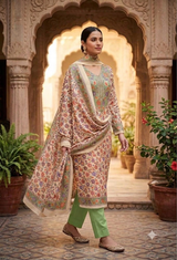 A woman models a traditional Indian salwar kameez suit in an ornate courtyard. The outfit features a beige kurta and dupatta with intricate multicolor floral embroidery, paired with solid green trousers and beige flat shoes.