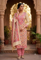 A woman stands in an ornate courtyard wearing a traditional Indian salwar kameez outfit. The outfit features a beige kurta with pink and orange floral embroidery and a matching dupatta, paired with pink trousers.