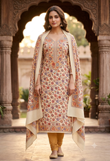 A woman models a traditional Indian salwar kameez suit set in front of an ornate stone archway. The set includes a beige kurta with a multicolor floral print, a matching beige dupatta, and mustard yellow trousers.