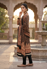 A woman stands in a stone courtyard wearing a long black and brown patterned kurta with ornate border details, paired with black pants and flat shoes, with arches and a fountain in the background.