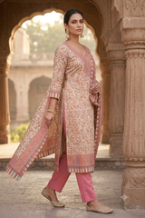 A woman models a traditional three-piece salwar kameez suit in a stone archway courtyard. The outfit features a light pink and beige floral print kurta with a matching printed dupatta, paired with solid pink trousers and beige flat shoes.