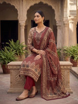 A model wearing an elegant deep maroon printed unstitched woolen suit featuring intricate beige patterns, a matching printed stole, and solid maroon bottoms, seated in a traditional courtyard.