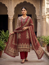 A model wearing an elegant deep maroon printed unstitched woolen suit featuring intricate beige patterns, a matching printed stole, and solid maroon bottoms, seated in a traditional courtyard.