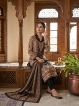 A woman sits on a carved wooden bench in a traditional interior, wearing a brown and black patterned ethnic outfit with a long draped dupatta. The setting features ornate wooden arches, windows, and a potted plant.