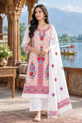 A woman models a traditional South Asian salwar kameez suit set outdoors. The set features a white kurta heavily embroidered with pink, red, and green floral patterns, paired with matching white trousers and a white dupatta decorated with pink and red floral motifs and a patterned border.
