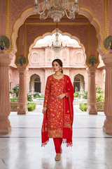 A woman modeling a red Indian ethnic salwar kameez suit with intricate gold and yellow floral embroidery, standing in a grand, ornate pink marble courtyard with arches and chandeliers.