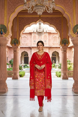 A woman stands in a grand pink stone courtyard with arches and chandeliers, wearing a bright red traditional outfit with gold floral embroidery and a matching dupatta.