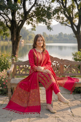 A woman wearing a bright pink and gold traditional South Asian outfit sits on a wooden bench outdoors by a lake, with trees and hills in the background.