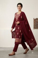 A woman models a traditional three-piece salwar kameez suit in deep maroon. The set features a long-sleeved kurta with colorful floral embroidery on the neckline and hem, matching trousers, and a coordinating dupatta draped over her shoulder.