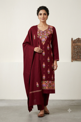 A woman models a traditional Indian salwar kameez suit in deep maroon with colorful floral embroidery, paired with a matching dupatta, set against a neutral indoor background.