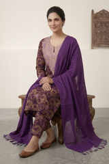 A woman seated on a wooden chair wearing a purple and brown patterned traditional outfit with a matching draped scarf, photographed in an indoor studio setting.