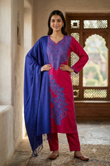 A woman models a traditional Indian salwar kameez suit with a vibrant magenta kurta featuring blue embroidery, matching magenta trousers, and a royal blue dupatta draped over her shoulder, set against a rustic indoor background.