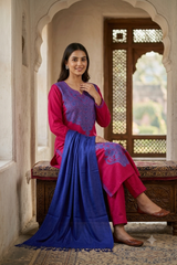 A woman models a traditional Indian salwar kameez suit in a courtyard setting. The outfit features a magenta-pink kurta with a purple-blue embroidered yoke, matching pink trousers, and a contrasting royal blue dupatta draped over a carved wooden bench.