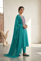 A woman models a teal green ethnic salwar kameez suit with a matching dupatta in a bright, modern room with neutral walls.