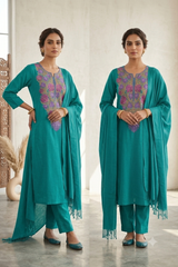 Two women modeling a teal green salwar kameez suit with purple and gold embroidery on the chest, paired with matching pants and a draped scarf, in a bright indoor setting.