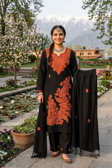 A woman stands outdoors in a garden courtyard wearing a black traditional outfit with prominent orange embroidery and holding a matching black dupatta. Snow-capped mountains, trees, and flowers are visible in the background.