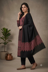 A woman models a black kurta set with deep red and gold border embroidery, paired with black pants and brown flat shoes, standing in front of a textured beige wall with a potted plant.