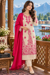 A woman models a traditional South Asian salwar kameez suit set on a wooden balcony with a scenic lake and snow-capped mountains in the background. The set features a cream-colored kurta with vibrant pink floral and peacock embroidery, matching cream trousers, and a bright pink dupatta.