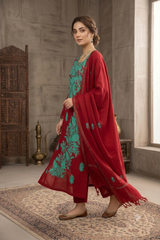 A woman models a traditional Indian outfit consisting of a red kurta with green floral embroidery, matching red trousers, and a red dupatta draped over her shoulder, set in a rustic room with a patterned rug.