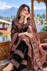 A woman sits on a wooden boat or dock railing in front of a scenic lake and snow-capped mountains, wearing a black traditional outfit with ornate gold embroidery and a dusty rose patterned dupatta.
